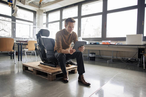 Ingenieur mit Tablet-PC auf einem Stuhl über einer Palette in der Werkstatt sitzend - JOSEF23626