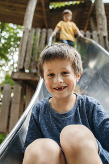 Glücklicher Junge rutscht auf der Rutsche mit seinem Bruder im Hintergrund auf dem Spielplatz hinunter - ELMF00004