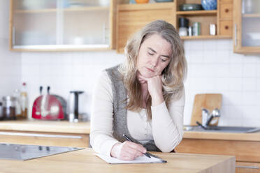 Reife Frau schreibt auf Papier in der Küche zu Hause - SGF02960