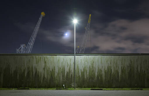 Netherlands, South Holland, Rotterdam, Old harbor cranes seen from behind surrounding wall at night - MKJF00034