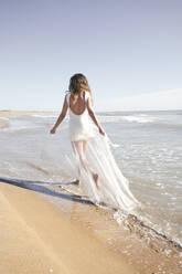 Junge Braut trägt weißes Hochzeitskleid und läuft am Strand - VEGF06254