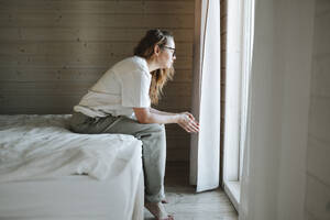 Besorgte Frau sitzt auf dem Bett und schaut durch das Fenster zu Hause - OLRF00200