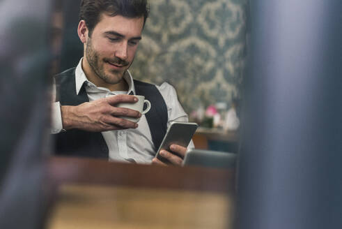 Geschäftsmann hält eine Kaffeetasse und benutzt ein Smartphone in einem Café - UUF31441