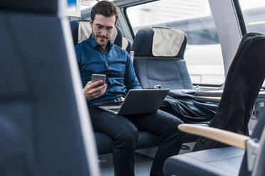 Businessman sitting with laptop and using smart phone in train - UUF31445