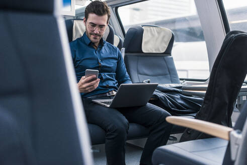 Geschäftsmann mit Laptop sitzend und mit Smartphone im Zug - UUF31445