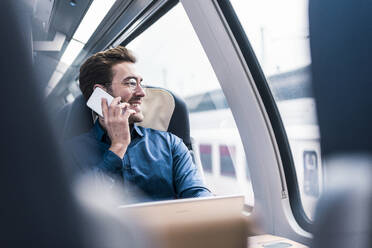 Lächelnder Geschäftsmann, der mit seinem Smartphone spricht und durch ein Fenster im Zug schaut - UUF31446