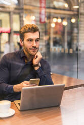 Young businessman sitting with smart phone and laptop in coffee shop - UUF31451