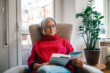 Mature woman sitting and reading book on armchair at home - GDBF00135