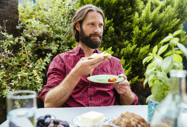 Mature man having sandwich at table in garden - MBEF01477