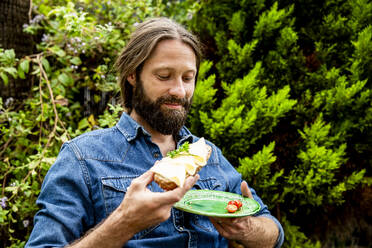 Mann mit Bart isst Käsesandwich im Garten - MBEF01478