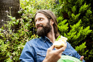Lächelnder Mann mit Bart hält Käsesandwich im Garten - MBEF01479