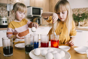 Sisters dipping Easter eggs in colored dye at home - ASHF00052