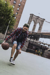 Young man playing basketball at court near Brooklyn Bridge in city - UUF31478