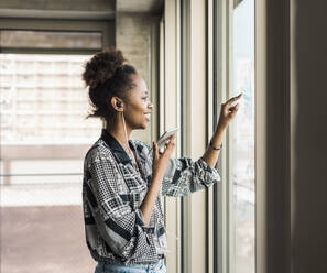 Young businesswoman with smart phone looking out window in office - UUF31500