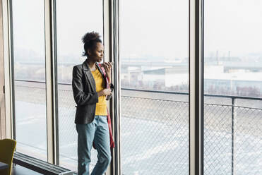 Junge Geschäftsfrau mit Mobiltelefon, die aus dem Fenster im Büro schaut - UUF31515