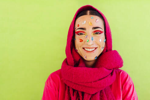Lächelnde Frau mit Edelsteinaufklebern im Gesicht und Schal vor grünem Hintergrund - EGHF00850