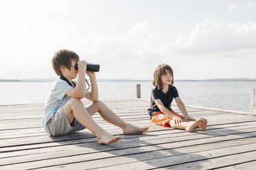 Junge schaut durch ein Fernglas und sitzt mit seinem Bruder auf einem Pier am See - ELMF00024