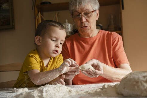 Senior woman teaching grandson to make dough buns in kitchen at home - KVBF00032