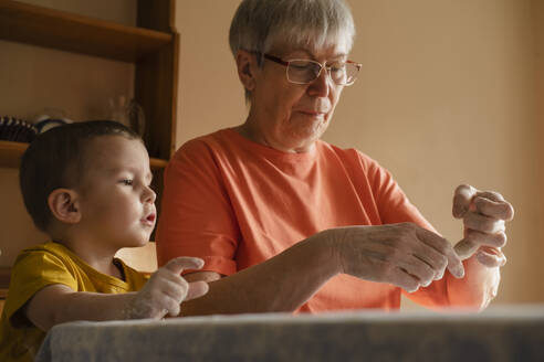 Senior woman preparing dough buns near grandson in kitchen at home - KVBF00034