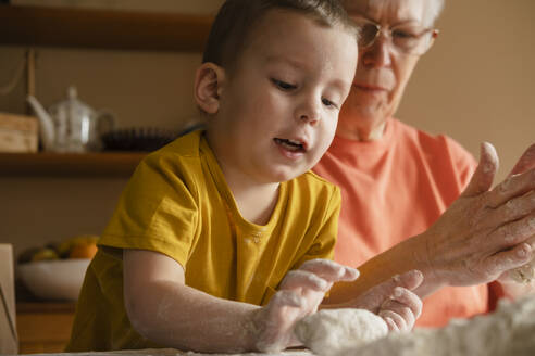 Boy helping grandmother in preparing dough buns in kitchen at home - KVBF00035