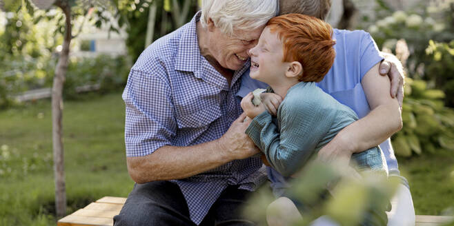 Cheerful boy having fun with grandparents sitting on bench in garden - MBLF00279