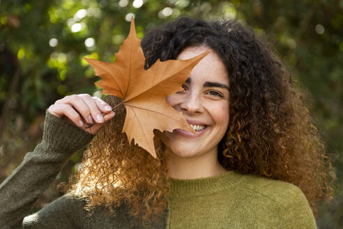 Smiling woman holding maple leaf near face at park - LMCF00930