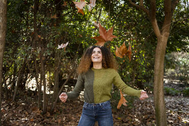 Playful woman standing under falling leaves at park - LMCF00932
