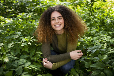 Lächelnde junge Frau, die inmitten von Pflanzen im Park kauert - LMCF00933
