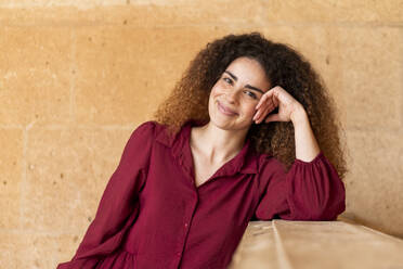 Smiling woman with curly hair leaning on elbow in front of wall - LMCF00959