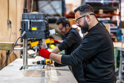 Worker operating electric drill machinery with colleague working in background - DLTSF03813