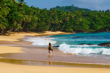 Back view of unrecognizable traveler strolls along the pristine Praia Emilia, with golden sands, lush palm trees, and turquoise waves touching the shore - ADSF52970