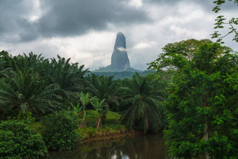 Serene landscape featuring the towering Pico Cão Grande amidst verdant tropical vegetation under moody skies, reflecting in a calm river. - ADSF52975