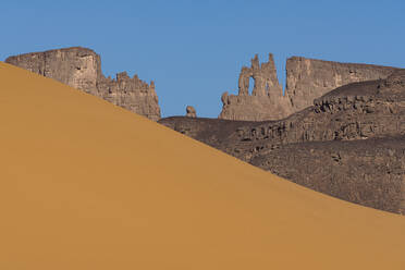 Scenic view of the Tadrart Rouge in Tin Merzouga landscape at dusk with golden sand dunes and rugged rock formations against a clear blue sky - ADSF52985