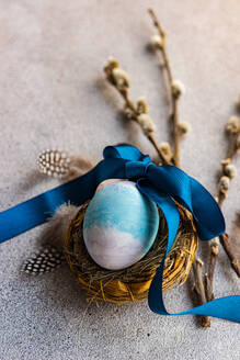 Nahaufnahme eines eleganten Oster-Flatlays mit einem blauen Ei in einem mit einem Band gebundenen Nest, begleitet von Weidenzweigen und Federn auf einer strukturierten Oberfläche - ADSF52994