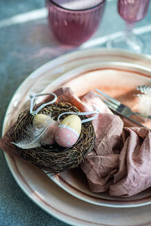 An artistic Easter table setting featuring a nest with pastel decorated eggs atop ceramic plates, complemented by a dusty pink napkin. - ADSF52998