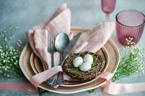 Eleganter Ostertisch mit pastellfarbenen Servietten, Tellern und dekorativen Eiern in einem Zweignest, ergänzt durch Blumen und Bänder. - ADSF52999