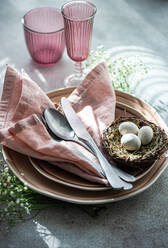 Eine elegante Osterdekoration mit einem Nest mit gesprenkelten Eiern, rosa Servietten und farblich passenden Gläsern auf einer strukturierten Oberfläche. - ADSF53000