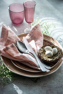 An elegant Easter dining setup featuring a nest with speckled eggs, pink napkins, and matching colored glassware on a textured surface. - ADSF53000