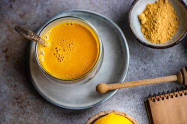 Ein warmer, goldener Kurkuma-Latte sitzt in einer Keramiktasse, begleitet von einer Schale Kurkumapulver und einem Honiglöffel, und vermittelt eine gemütliche, gesunde Atmosphäre - ADSF53009