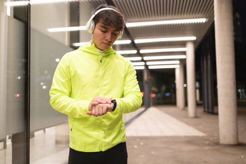Seriöser junger Mann in gelber Jacke mit Kopfhörern, der die Zeit auf seiner Armbanduhr überprüft, während er in der Nähe der Glaswand eines beleuchteten modernen Gebäudes in der Nacht steht - ADSF53170