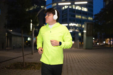 Nachdenklicher junger Mann in legerer Sportkleidung und kabellosen Kopfhörern, der Musik hört und wegschaut, während er nachts auf einer beleuchteten Stadtstraße mit gepflastertem Boden joggt - ADSF53172