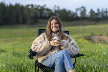 Smiling young woman holding coffee cup sitting on camping chair - JSIF00007