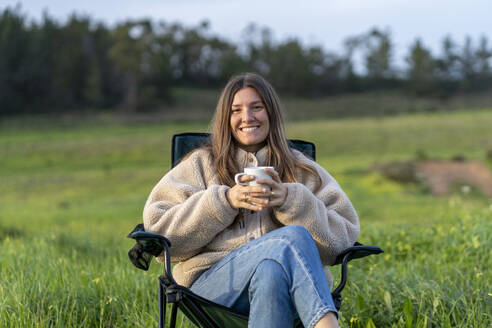 Lächelnde junge Frau mit Kaffeetasse auf einem Campingstuhl sitzend - JSIF00007