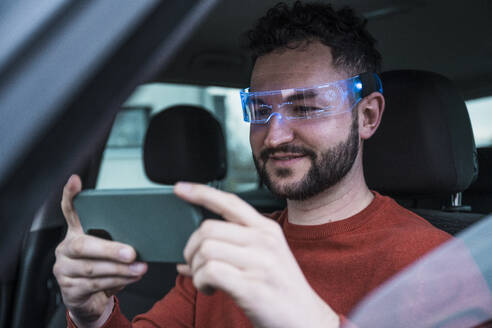 Smiling man wearing smart glasses and using smart phone in car - UUF31562