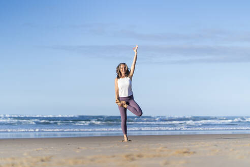 Ältere Frau macht Baum-Yoga-Pose mit erhobener Hand am Strand - JSIF00017