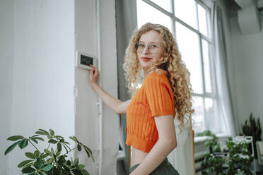 Smiling woman with blond hair standing near thermostat on wall at home - MDOF01852