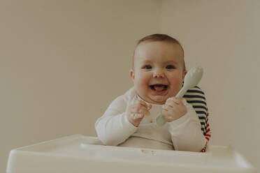 Happy toddler holding spoon and sitting on chair at home - OSF02435