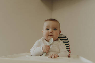 Baby boy holding spoon and sitting on chair at home - OSF02436