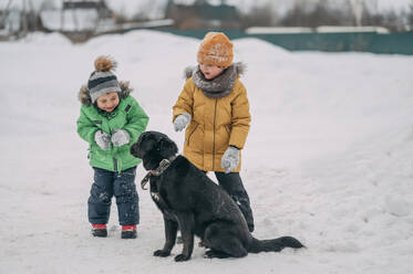 Glückliche Kinder in warmer Kleidung und beim Spielen mit dem Hund im Schnee - ANAF02729