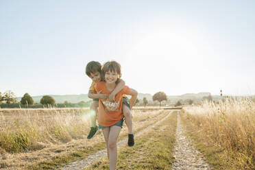 Smiling girl piggybacking brother in field at sunset - ELMF00027